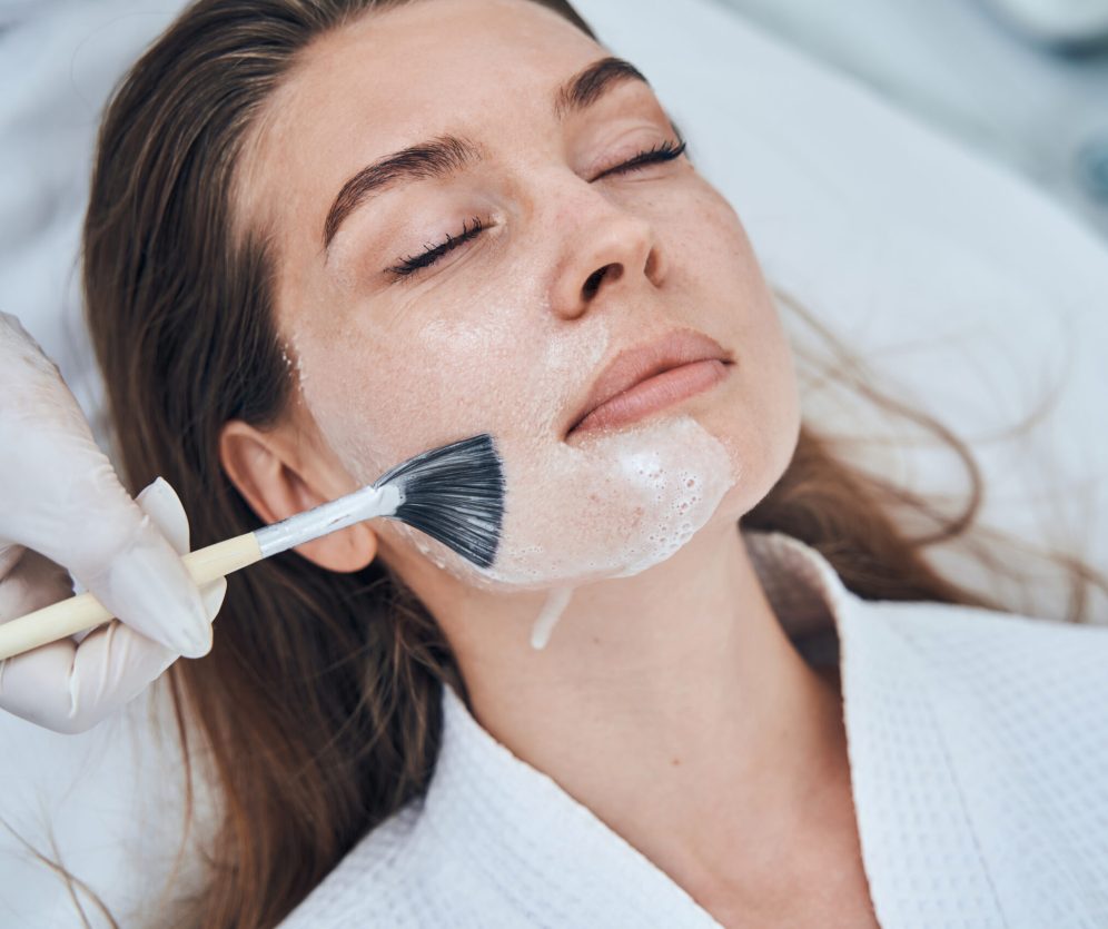 Competent cosmetologist wearing rubber gloves while using brush for applying mask on face